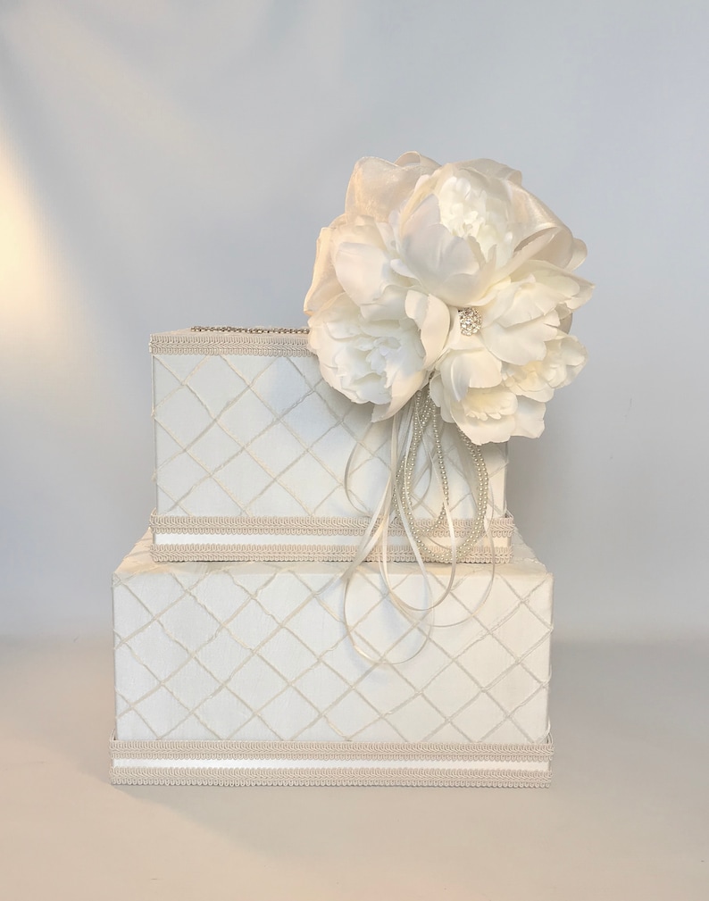 May include: White wedding card box with a white flower on top. The box has a diamond pattern and a beige ribbon around the top and bottom.