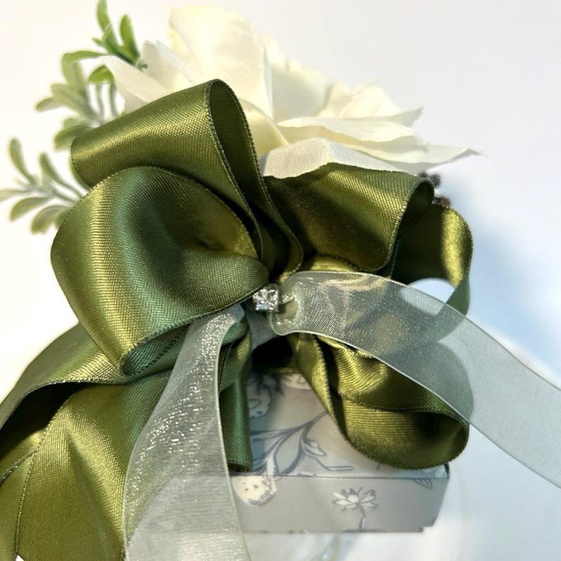 K&ouml;nnte beinhalten: Eine kleine Geschenkbox, die in ein floral gemustertes Papier eingewickelt ist, mit einem gr&uuml;nen Satinband, das zu einer Schleife gebunden ist. Die Schleife ist mit einem klaren Strassstein und einem durchsichtigen gr&uuml;nen Band verziert.