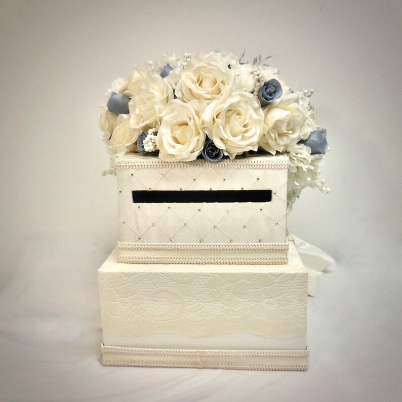 Pu&ograve; includere: Una scatola per biglietti di nozze bianca a due livelli con un arrangiamento floreale bianco e blu in cima. Il livello superiore ha una fessura per i biglietti ed &egrave; decorato con un motivo a rombi. Il livello inferiore ha un motivo a pizzo.