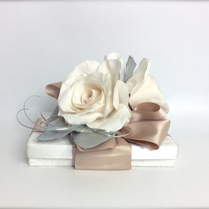 May include: A white gift box with a beige satin ribbon and a white rose on top. The box is wrapped in white paper and has a silver leaf design.