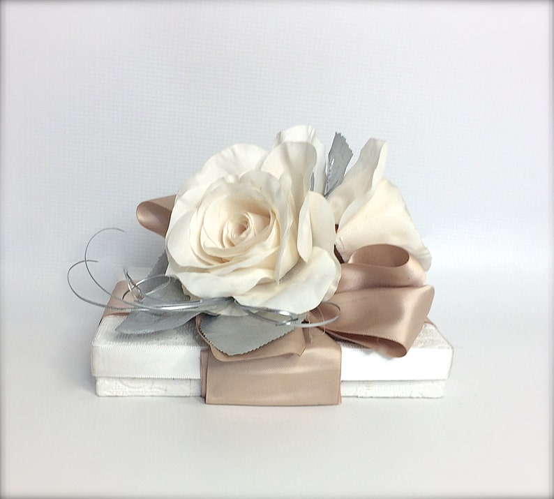 May include: A white gift box with a beige satin ribbon and a white rose on top. The box is wrapped in white paper and has a silver leaf design.