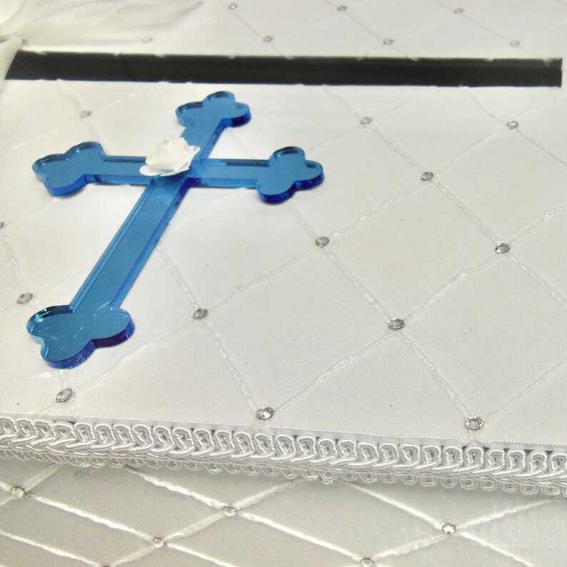 May include: White satin box with a blue cross on top. The box has a white trim around the edge and a diamond pattern on the surface.