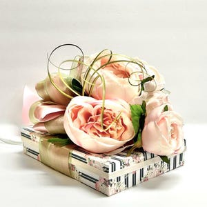 K&ouml;nnte beinhalten: Eine dekorative Geschenkbox mit floralem und gestreiftem Muster, gekr&ouml;nt von hellrosa Pfingstrosen und einer gro&szlig;en Schleife. Die Schleife besteht aus rosa und goldenem Band mit gr&uuml;nen Akzenten. Die Box ist mit einem goldenen Band umwickelt.