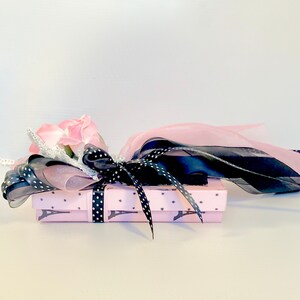 K&ouml;nnte beinhalten: Eine kleine rosa Geschenkbox mit einem schwarz-wei&szlig;en Polka-Dot-Band und einem rosa Eiffelturm-Muster. Die Box ist mit einer rosa Rose und schwarzen und rosa B&auml;ndern verziert.