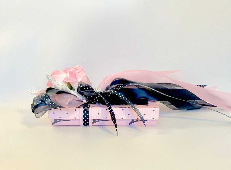 K&ouml;nnte beinhalten: Eine kleine rosa Geschenkbox mit einem schwarz-wei&szlig;en Polka-Dot-Band und einem rosa Eiffelturm-Muster. Die Box ist mit einer rosa Rose und schwarzen und rosa B&auml;ndern verziert.