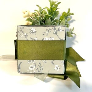 K&ouml;nnte beinhalten: Eine quadratische Geschenkbox mit einem Blumenmuster und einem gr&uuml;nen Band. Die Box ist mit einer kleinen Topfpflanze gef&uuml;llt.