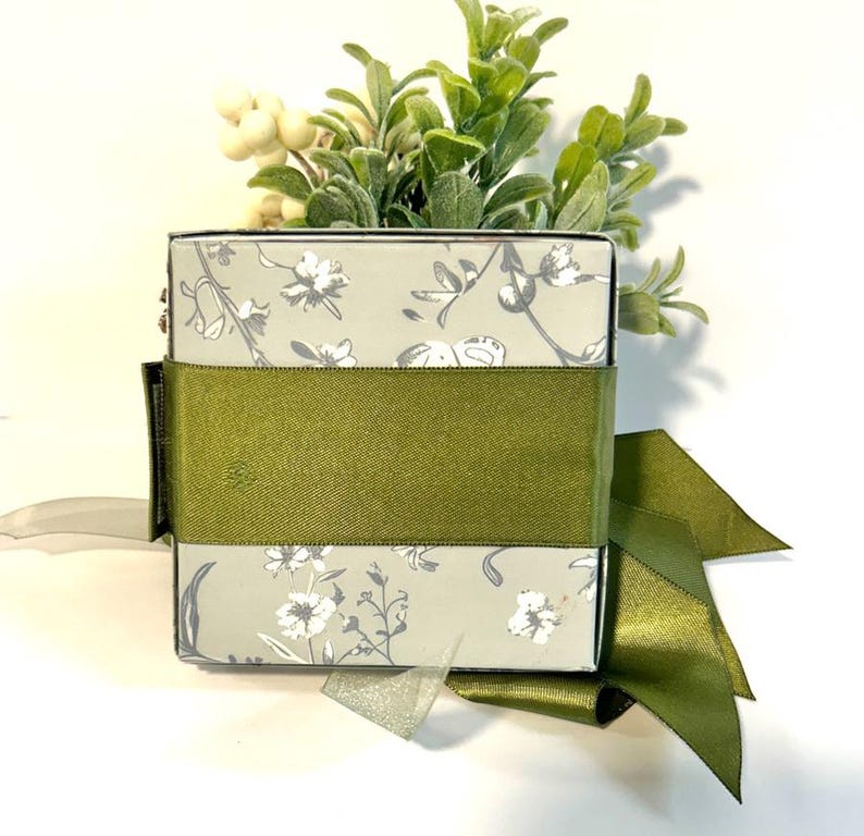 K&ouml;nnte beinhalten: Eine quadratische Geschenkbox mit einem Blumenmuster und einem gr&uuml;nen Band. Die Box ist mit einer kleinen Topfpflanze gef&uuml;llt.