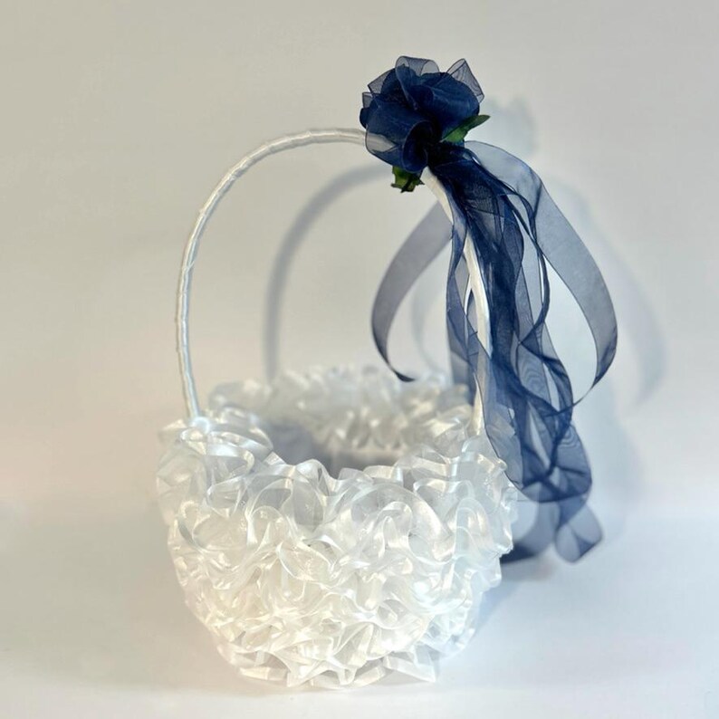 May include: A white flower girl basket with a ruffled design and a handle. The basket is decorated with a navy blue flower and sheer ribbons. The basket is designed for a wedding.