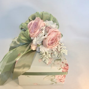 Puede incluir: Una caja de regalo blanca con un dise&ntilde;o floral y una cinta de sat&eacute;n verde atada con un lazo. El lazo est&aacute; adornado con rosas rosas y flores blancas.