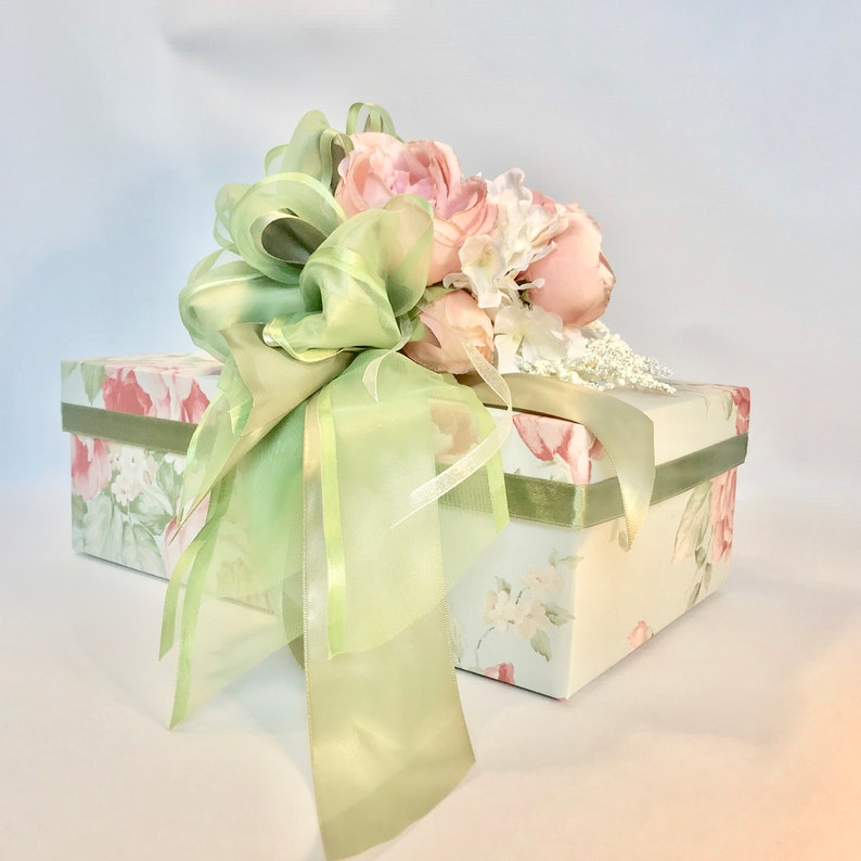 Puede incluir: Una caja de regalo blanca con un patr&oacute;n floral y una cinta verde atada en un lazo. La caja est&aacute; coronada con un ramo de flores rosas y blancas.