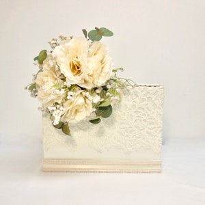 May include: White wedding card box with a lace design and a bouquet of white roses and greenery on top.