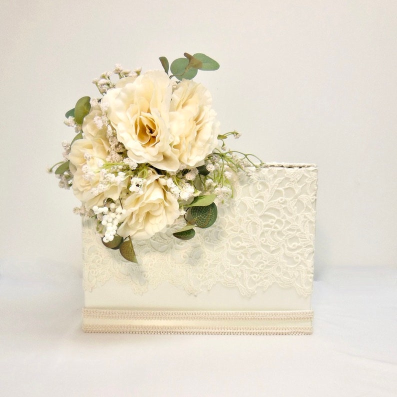 May include: White wedding card box with a lace design and a bouquet of white roses and greenery on top.