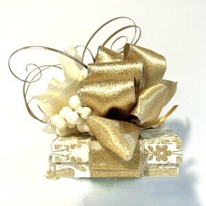 May include: A small white gift box with a gold ribbon and bow. The box is decorated with a floral pattern and has a white flower and pearl embellishment on top.