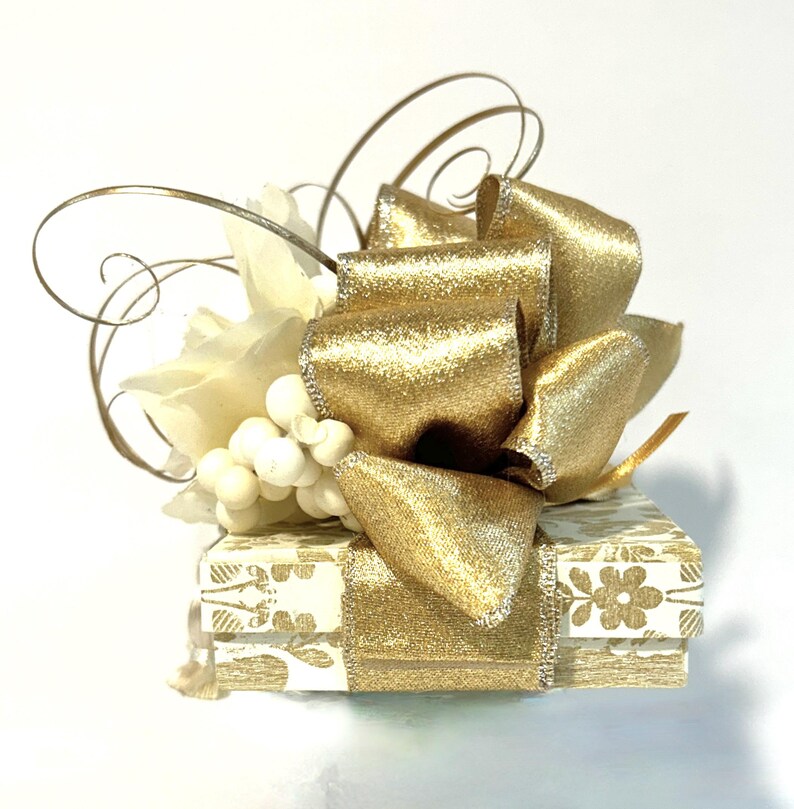 May include: A small white gift box with a gold ribbon and bow. The box is decorated with a floral pattern and has a white flower and pearl embellishment on top.