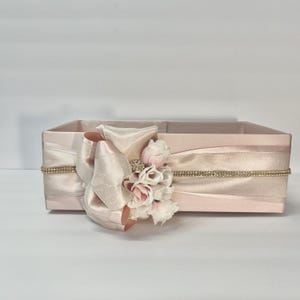 May include: A rectangular, light pink box with a large, cream-colored bow and floral arrangement. The box is adorned with a thin, gold-colored rhinestone trim. The bow and flowers add a touch of elegance to this decorative storage container.