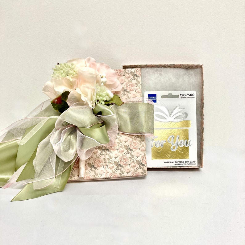 May include: A gift box wrapped in a floral patterned paper with a green ribbon and a bouquet of pink and white flowers. The box is next to a gift card with the text "For You" and "American Express Gift Card".