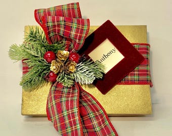May include: A rectangular gold gift box wrapped with a red, green, and gold plaid ribbon. The ribbon is tied in a bow and decorated with artificial pine needles, red berries, and gold flowers. A square maroon velvet tag displays the name Anthony.