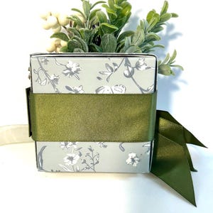 K&ouml;nnte beinhalten: Eine kleine, quadratische Geschenkbox mit einem Blumenmuster in Graut&ouml;nen und Wei&szlig;. Die Box ist mit einem gr&uuml;nen Band und einer Schleife verziert.