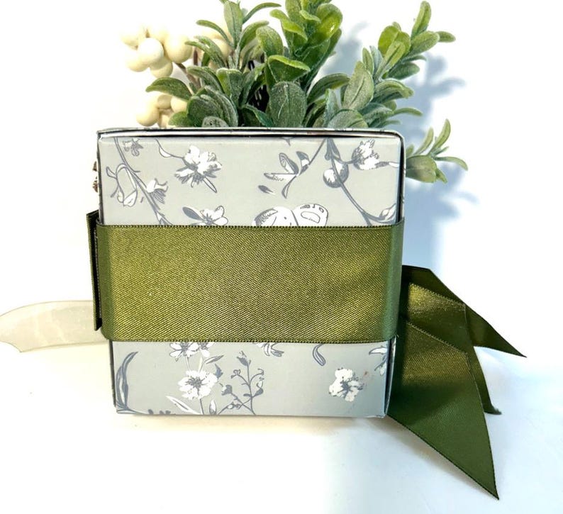 K&ouml;nnte beinhalten: Eine kleine, quadratische Geschenkbox mit einem Blumenmuster in Graut&ouml;nen und Wei&szlig;. Die Box ist mit einem gr&uuml;nen Band und einer Schleife verziert.