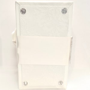 May include: A white, rectangular wedding card box with a floral embossed design. The box is adorned with four sparkling, round rhinestone embellishments. A white satin ribbon wraps around the center.