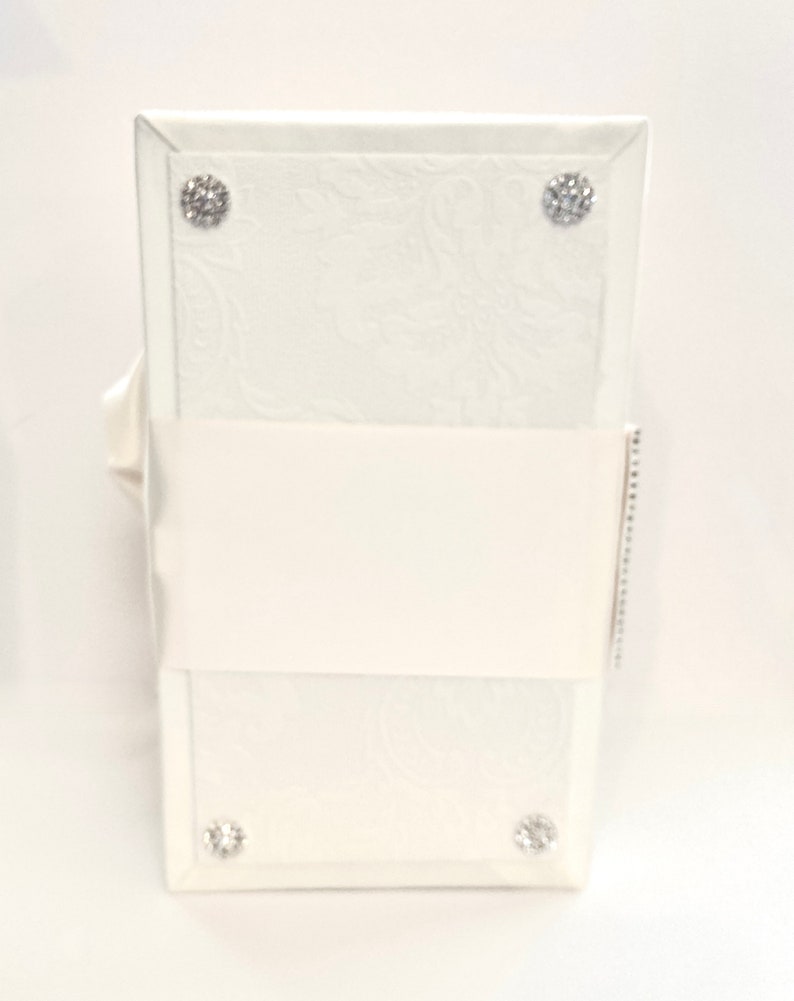 May include: A white, rectangular wedding card box with a floral embossed design. The box is adorned with four sparkling, round rhinestone embellishments. A white satin ribbon wraps around the center.