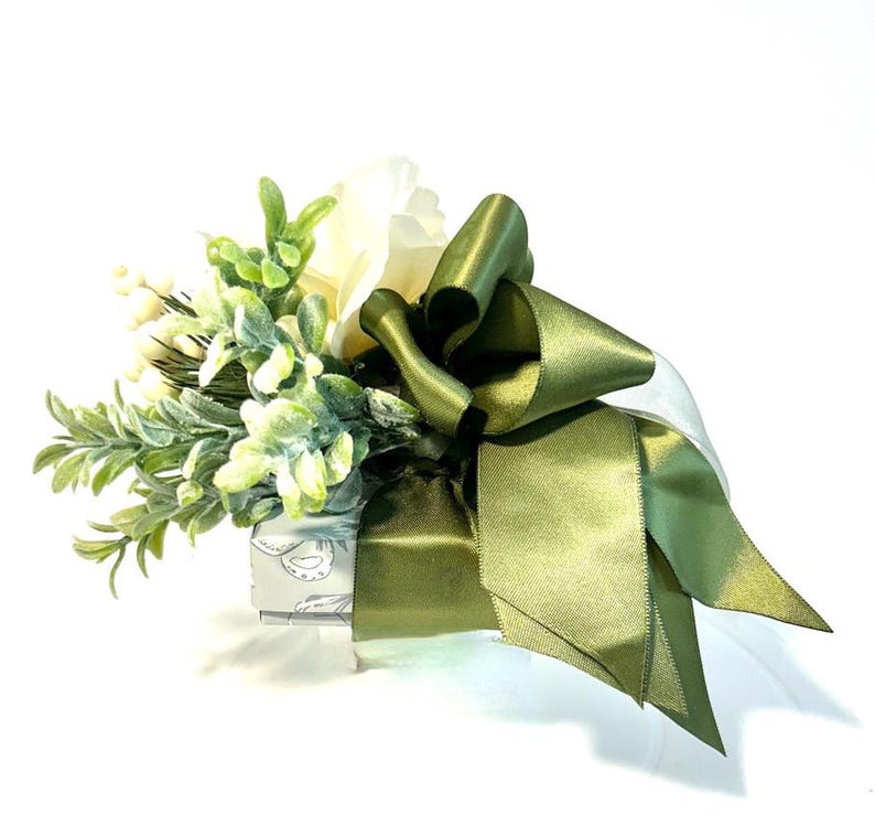 K&ouml;nnte beinhalten: Eine kleine Geschenkbox, die in wei&szlig;es Papier mit gr&uuml;nem Blumendesign eingewickelt ist. Die Box ist mit einem Strau&szlig; wei&szlig;er Blumen und gr&uuml;nem Laub verziert, das mit einer gro&szlig;en gr&uuml;nen Schleife aus Satin gebunden ist.