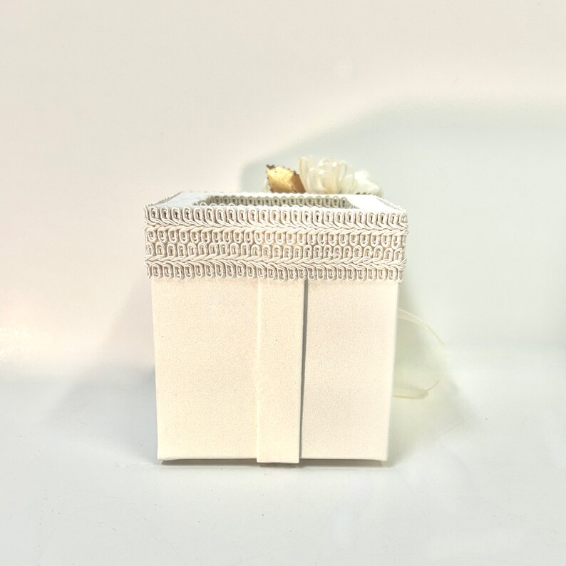 May include: A white gift box with a decorative trim around the top. The box is tied with a white ribbon and has a small white flower on top.