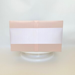 May include: A rectangular, blush-colored box wrapped with a wide white ribbon. The box is presented on a white, round pedestal, creating a clean and elegant aesthetic. The overall presentation suggests a gift or decorative item.