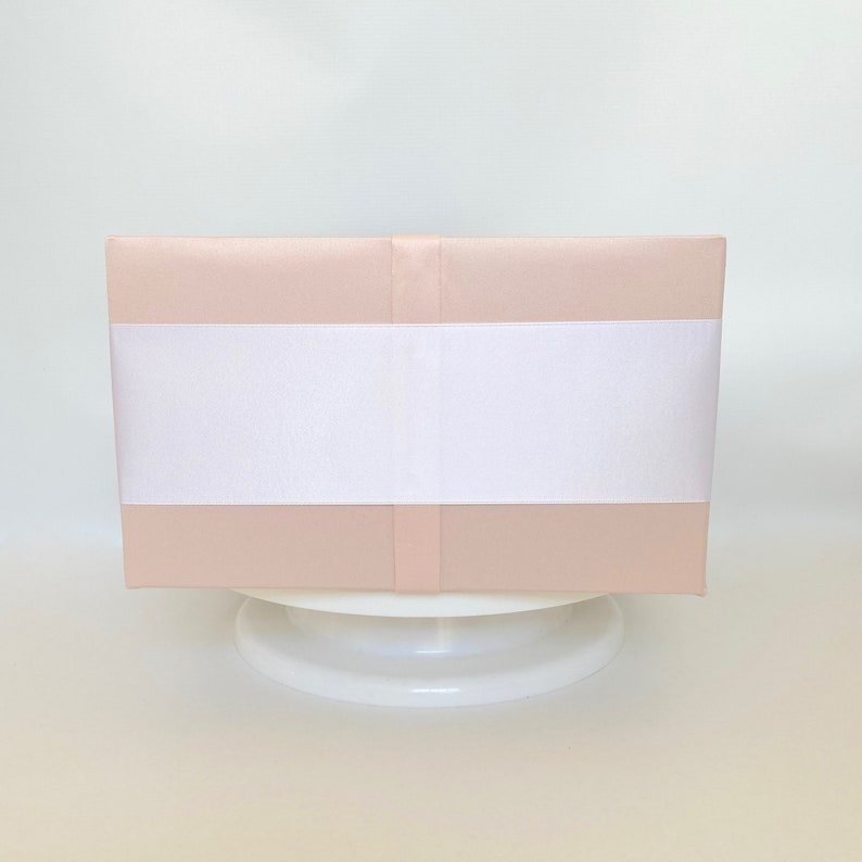 May include: A rectangular, blush-colored box wrapped with a wide white ribbon. The box is presented on a white, round pedestal, creating a clean and elegant aesthetic. The overall presentation suggests a gift or decorative item.