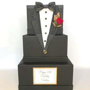 May include: A black tuxedo-shaped gift box with a red rose boutonniere, gold buttons, and a white bow tie. The box has a label that reads "Happy 50th Birthday Anthony".