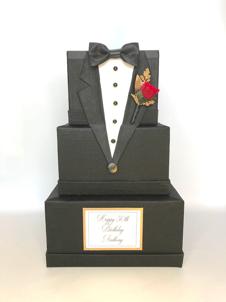 May include: A black tuxedo-shaped gift box with a red rose boutonniere, gold buttons, and a white bow tie. The box has a label that reads "Happy 50th Birthday Anthony".