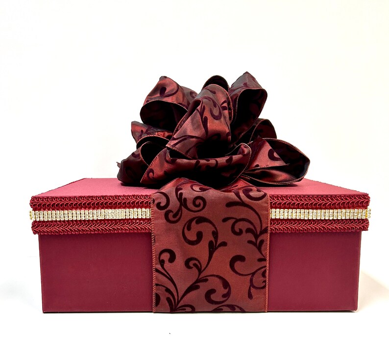 May include: A burgundy gift box with a large, ornate bow. The box is rectangular with a decorative trim of gold and rhinestones. A burgundy ribbon with a dark floral pattern wraps around the box.