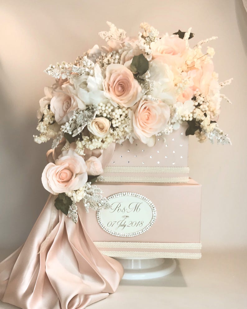 May include: Two-tiered pink wedding card box with a white satin bow and a floral arrangement of white and blush roses, white hydrangeas, and white berries. The box has a silver oval label with the text "Mr & Mrs 07 July 2018".