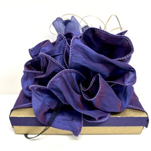 May include: A purple gift box with a large, ruffled purple bow. The bow is made of sheer fabric with a satin finish.