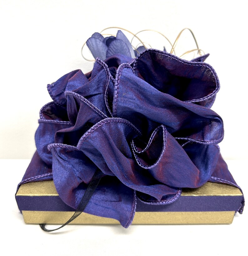 May include: A purple gift box with a large, ruffled purple bow. The bow is made of sheer fabric with a satin finish.