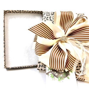 May include: A gift box with a leopard print design, open to reveal a brown and white striped bow with a rhinestone embellishment. The box is filled with white tissue paper.