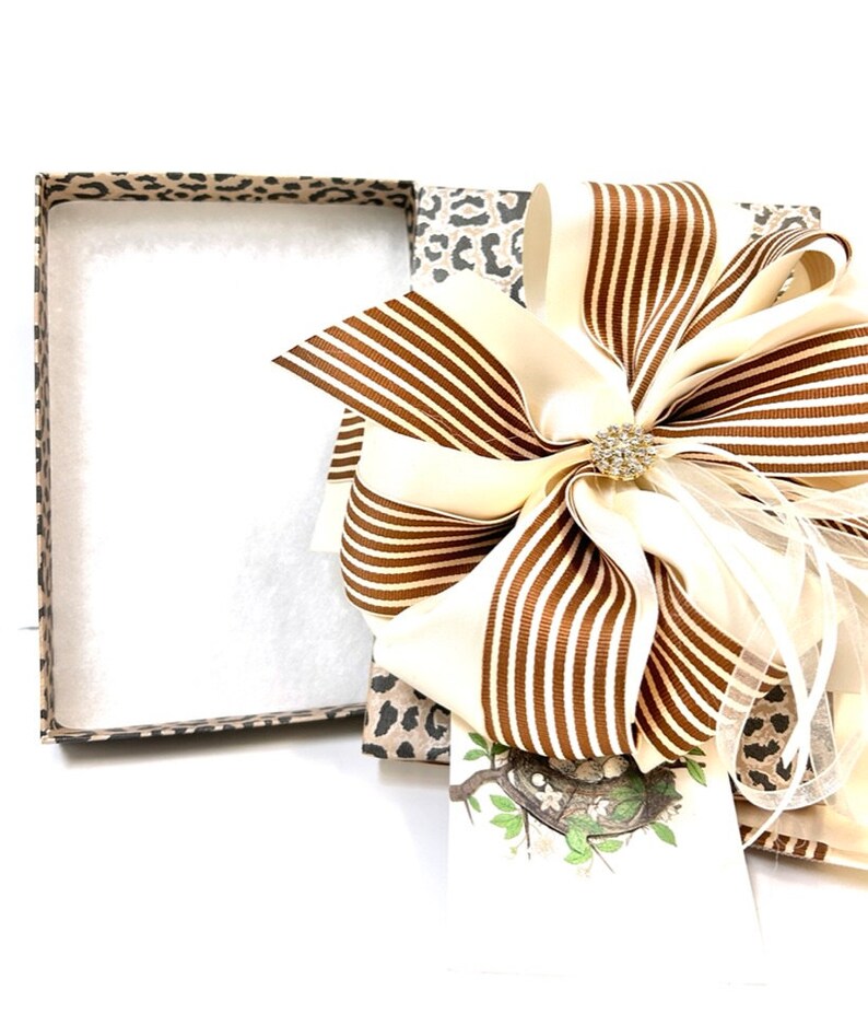 May include: A gift box with a leopard print design, open to reveal a brown and white striped bow with a rhinestone embellishment. The box is filled with white tissue paper.