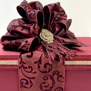 May include: A burgundy gift box with a large, ornate bow. The bow features a patterned design and is adorned with a jeweled brooch and tassel. The box is trimmed with rhinestone accents, creating an elegant presentation.