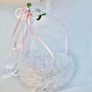 May include: White flower girl basket with a pink satin ribbon and white tulle. The basket is decorated with a white flower with a rhinestone center.