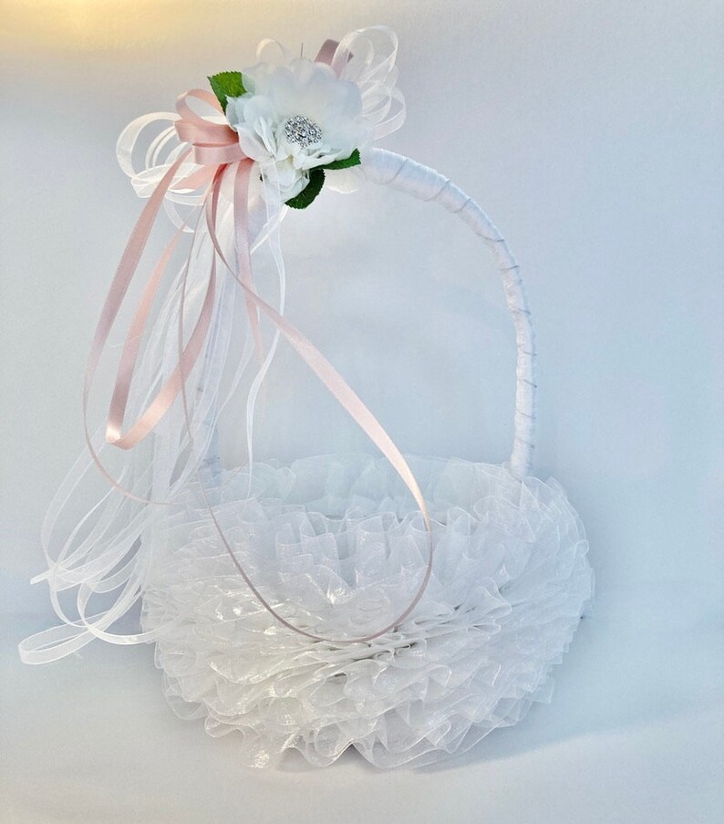 May include: White flower girl basket with a pink satin ribbon and white tulle. The basket is decorated with a white flower with a rhinestone center.