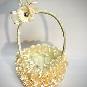 Peut inclure: Un panier de demoiselle d'honneur cr&egrave;me avec un design &agrave; volants. Le panier est dot&eacute; d'une poign&eacute;e et est orn&eacute; d'une grande fleur et d'un n&oelig;ud. La fleur a un centre en strass. Le panier est fait d'un mat&eacute;riau doux et soyeux.