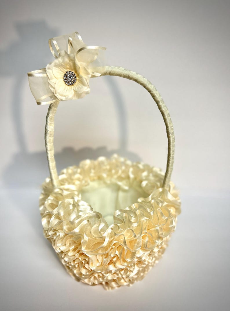Peut inclure: Un panier de demoiselle d'honneur cr&egrave;me avec un design &agrave; volants. Le panier est dot&eacute; d'une poign&eacute;e et est orn&eacute; d'une grande fleur et d'un n&oelig;ud. La fleur a un centre en strass. Le panier est fait d'un mat&eacute;riau doux et soyeux.