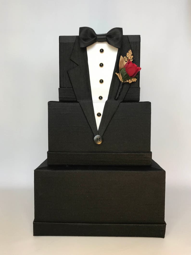 May include: A three-tiered black gift box set with a tuxedo design. The top tier features a black bow tie, white shirt, and a red rose buttonhole.