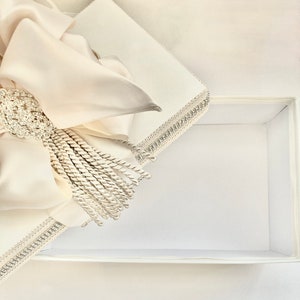 May include: An open, white rectangular gift box with a satin bow and tassel. The bow is cream-colored and adorned with a sparkling, silver-toned brooch. The box's edges are trimmed with silver-toned beading.