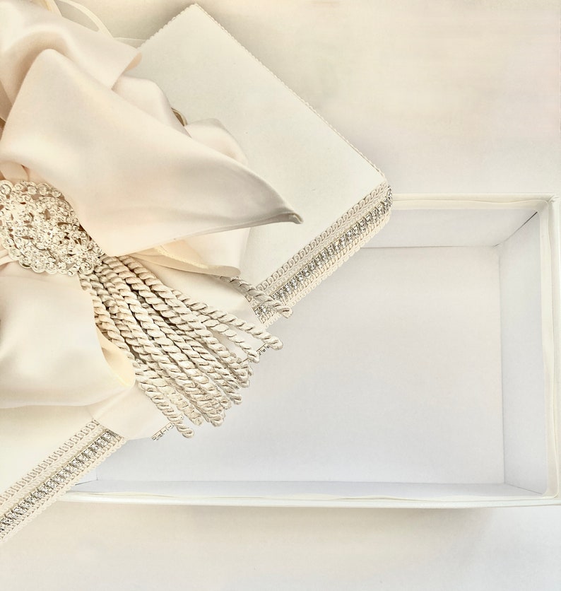 May include: An open, white rectangular gift box with a satin bow and tassel. The bow is cream-colored and adorned with a sparkling, silver-toned brooch. The box's edges are trimmed with silver-toned beading.
