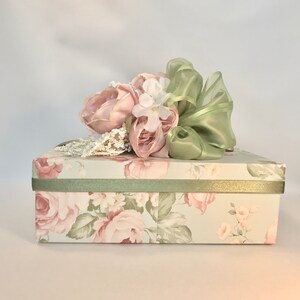 Puede incluir: Una caja de regalo floral con una cinta verde y un ramo de flores rosas y blancas.