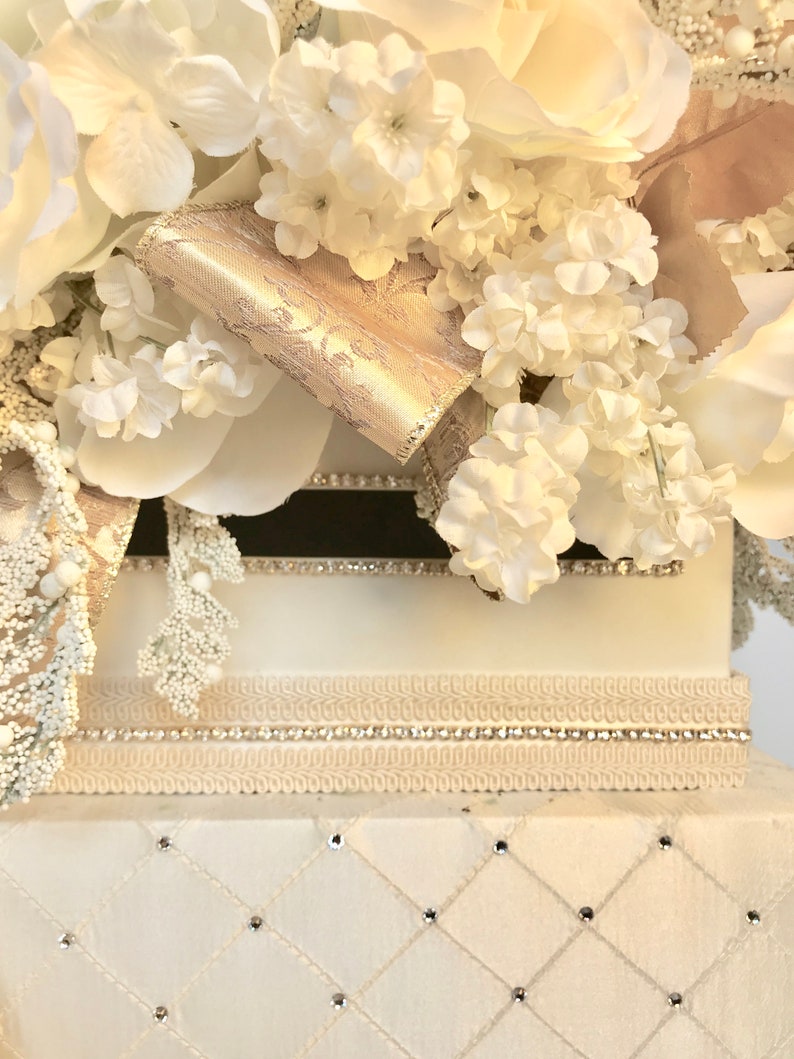 May include: White wedding card box with a white and gold floral arrangement on top. The box has a white satin base with a diamond pattern and rhinestones.