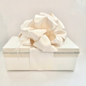 May include: A white gift box with a large ivory satin bow on top. The box is decorated with a silver and crystal trim around the top edge and a satin ribbon. The overall theme is elegant and suitable for special occasions.