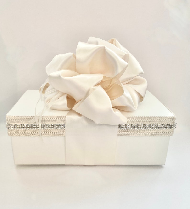 May include: A white gift box with a large ivory satin bow on top. The box is decorated with a silver and crystal trim around the top edge and a satin ribbon. The overall theme is elegant and suitable for special occasions.