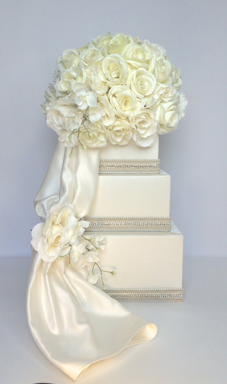 May include: A white tiered box with a satin ribbon and a bouquet of white roses and hydrangeas on top. The box is decorated with rhinestones.
